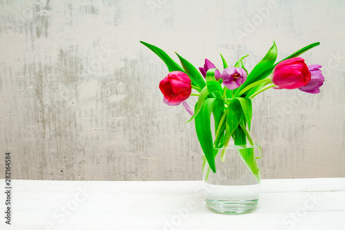 Copy space with beautiful pink and purple tulips with green leaves bouquet in a transparent glass vase on white wooden table on gray background