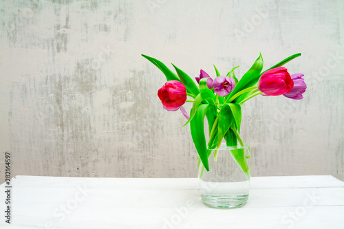 Copy space with beautiful pink and purple tulips with green leaves bouquet in a transparent glass vase on white wooden table on gray background