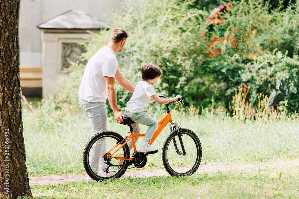 full length view of son riding bicycle and father walking next to kid and helping boy to ride on bike