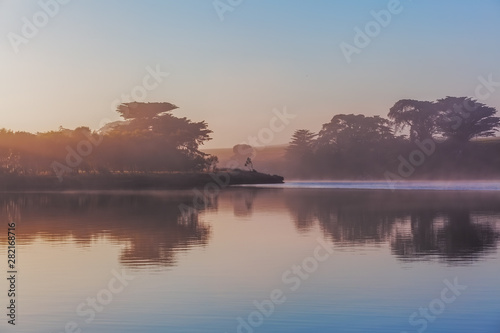 Trees silhouettes and early morning fog over river at sunrise