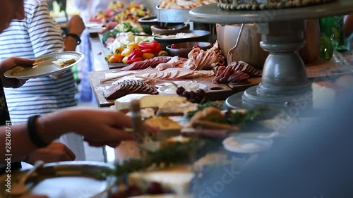 Smooth shot of a line of people at a crowded event going through a food line and grabbing a variety of food including meats, cheeses, bread, fruits, and vegetables. photo
