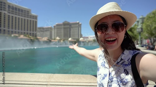 Young beautiful girl traveler standing in city near fountains outdoor summer. happy female backpacker waving hands while talking on video phone call. woman tourist showing camera view in background