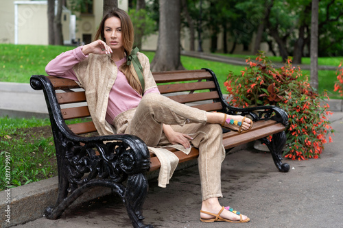 young European girl, natural appearance, long hair, beautiful face, coffee colored trousers and jacket, pink shirt to sit on wooden bench in park. Comfortable linen women's clothing. © Денис Бухлаев