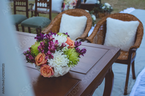 wooden table with a bouquet of colorful flowers and wooden chairs with white cushions on a gray carpet. Outdoor wedding ceremony.