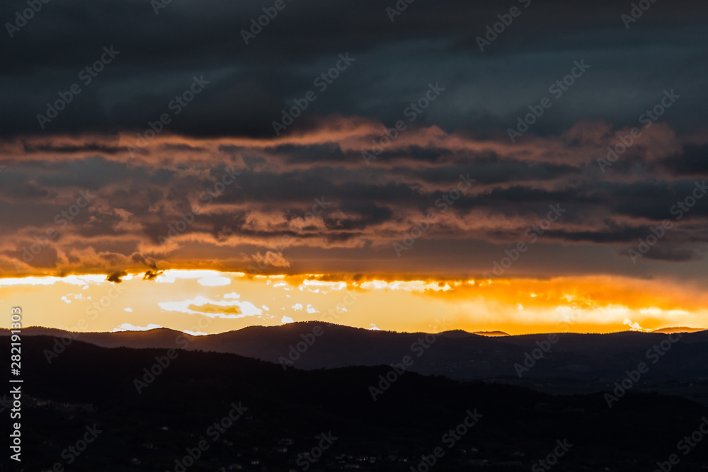 Fototapeta premium View of dark clouds at sunset, with sun filtering through an hole over the mountains