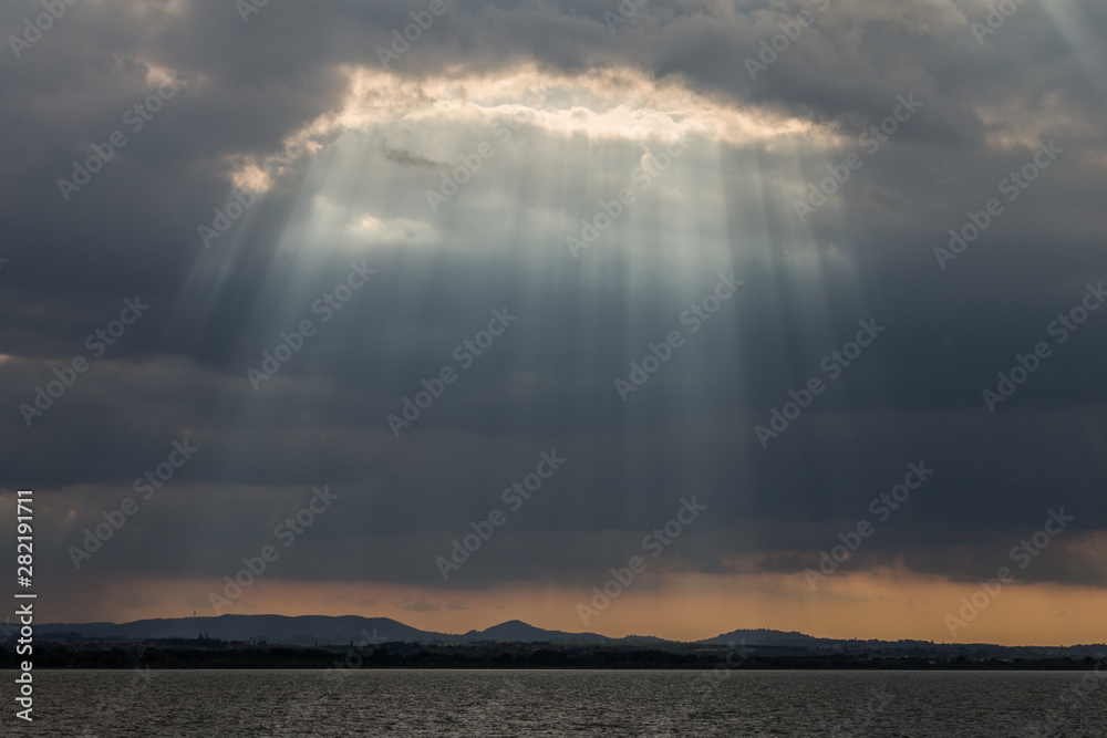 Sunrays at near sunset, with dark clouds in the background, an orange sky, and Trasimeno lake (Umbria, Italy) below