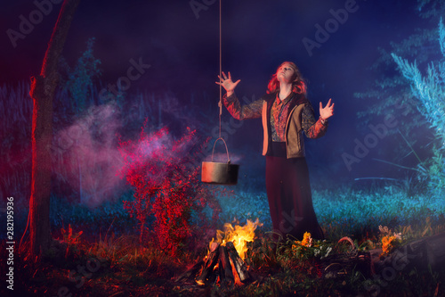 young witch by fire in night forest prepares magic potion