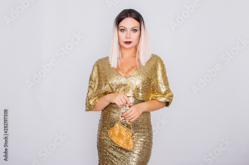 Charming young blonde haired woman wearing elegant gold evening dress and holding gold color clutch in her hands