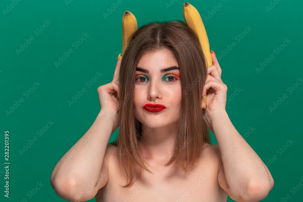 Closeup portrait of beautiful young woman with bright color make-up holding yellow bananas like ears standing isolated infront of a green background. Closeup portrait of beautiful young woman with bright color make-up holding yellow bananas like ears standing isolated infront of a green background.