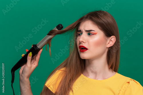 Portrait of beautiful young woman with bright color make-up having probems doing hair style herself with a curling iron. photo