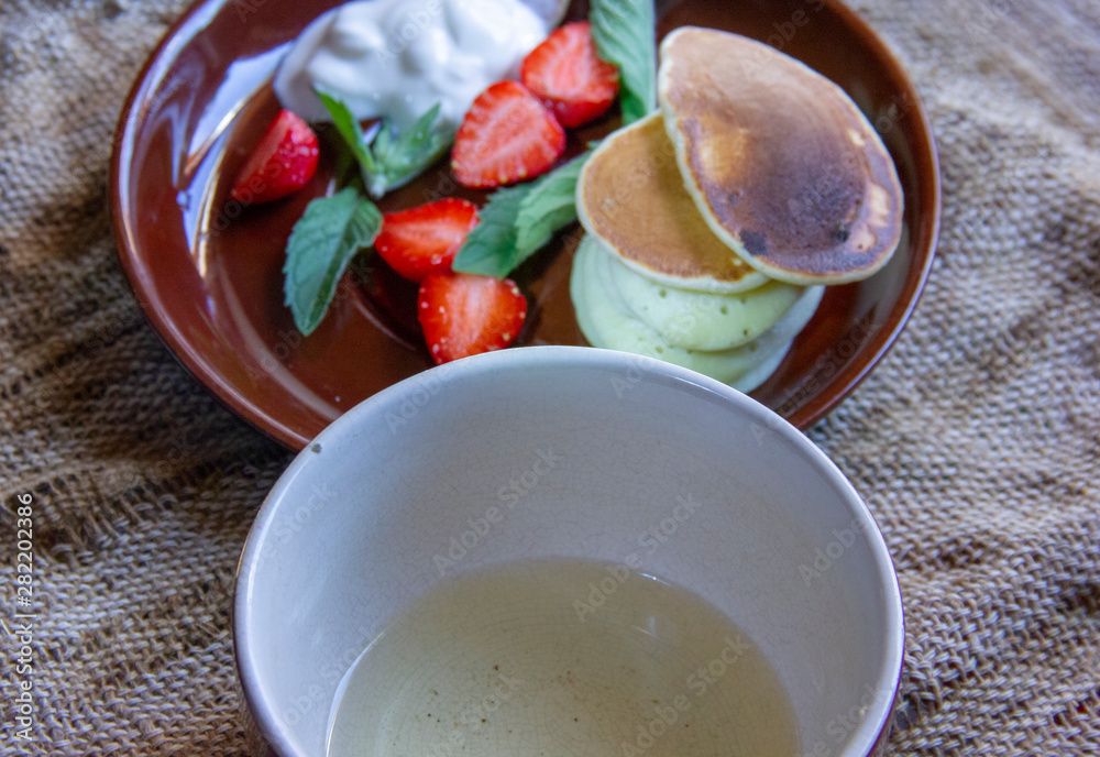 great breakfast. Fritters on plates with chopped strawberries and mint leaves. A plate and a cup of tea are on a jute napkin. great breakfast. Fritters on plates with chopped strawberries and mint leaves. A plate and a cup of tea are on a jute napkin.