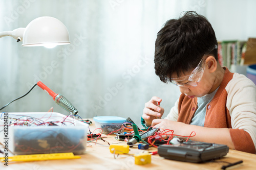 Smart preteen / teenage Asian boy screwdrivering, assembling and fixing computer chip, electronics hardware and circuits for school project with concentration and determination on desk. STEM Education