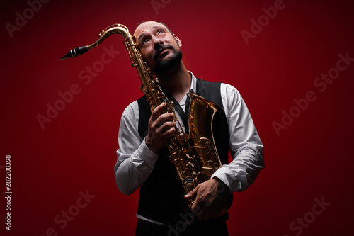 Portrait of professional musician saxophonist man in suit plays jazz music on saxophone, red background in a photo studio