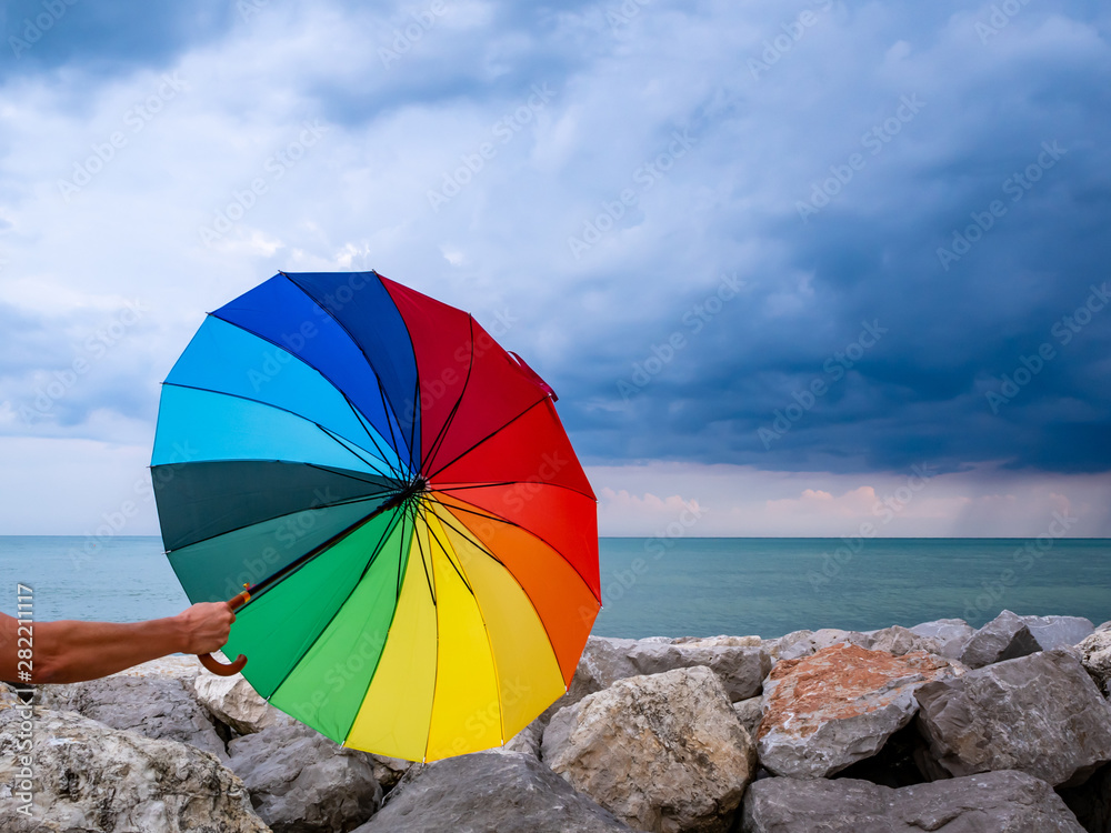 Männerhand mit bunten Regenschirm in der Hand am Meer, dunkle Regenwolken, Sturm
