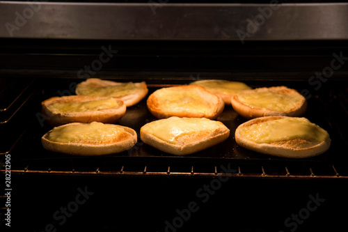 Slices of mild cheddar cheese under the grill for home made family burger night. Cheese is yellow, bread rolls are white and oven is dark with stainless steel edges. Hamburgers are a family favourite.