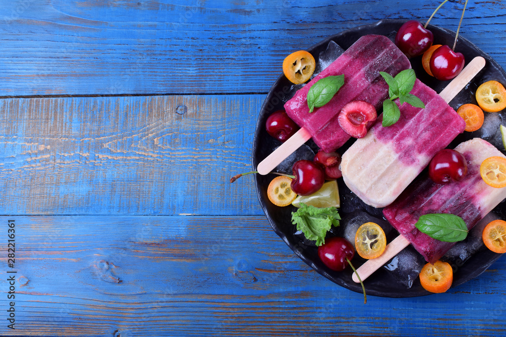 Fruit and berry popsicles on the plate with ice, pieces of kumquat, lime and cherries and mint. Copy space on blue table
