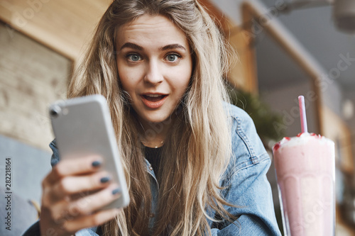 Girl cannot believe she received invitation on most famout summer party, sitting in cafe with friend, staring with popped eyes, dropping jaw from amazement, holding smartphone, reading impressive news photo