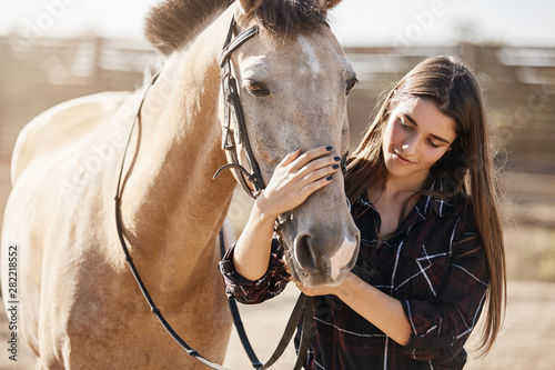 Animals, friendship and hobby concept. Pretty young woman in rural area smiling, gently petting horse nose, vet taking care of domestic pets on countryside farm, teaching children become jokey © Liubov Levytska