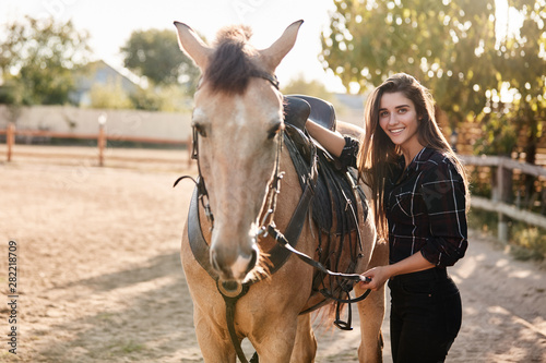 Girl prepare her horse for ride on racetrack. Cheerful female jockey fasten saddle, grooming cute animal and smiling joyfully camera, enjoy fresh air, taking care rancho, farm pets