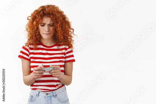 Tensed attractive redhead girl ginger female gaming geek pursing lips eager win rase playing smartphone game holding gadget look device display perplexed trying solve hard level, white background