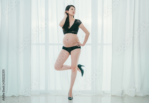 Full body portrait of sexy pregnant female model with long black hair wearing black lace bra and underwear standing leaning against window and posing. hot future parenthood woman in high heeled shoes © PR Image Factory
