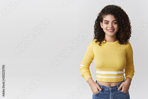 Attractive stylish millennial young hispanic girl smiling friendly hold hands jeans pockets grinning delighted standing have conversation campus hallway talking friend casually white background