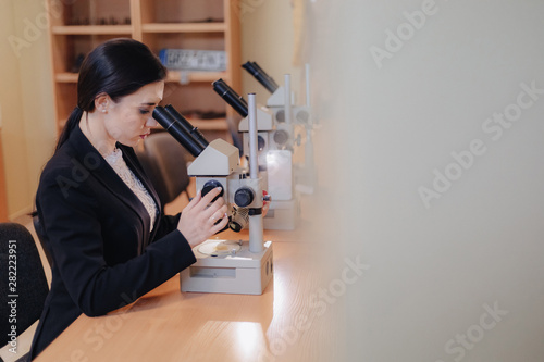 Young emotional attractive girl sitting at the table and working with a microscope in a modern office or audience