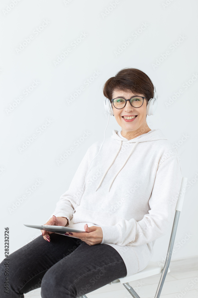 Charming slim middle-aged woman in glasses and a sweater sits on a chair and listens to her favorite online radio with the help of a headset and wireless internet and holding tablet on a white