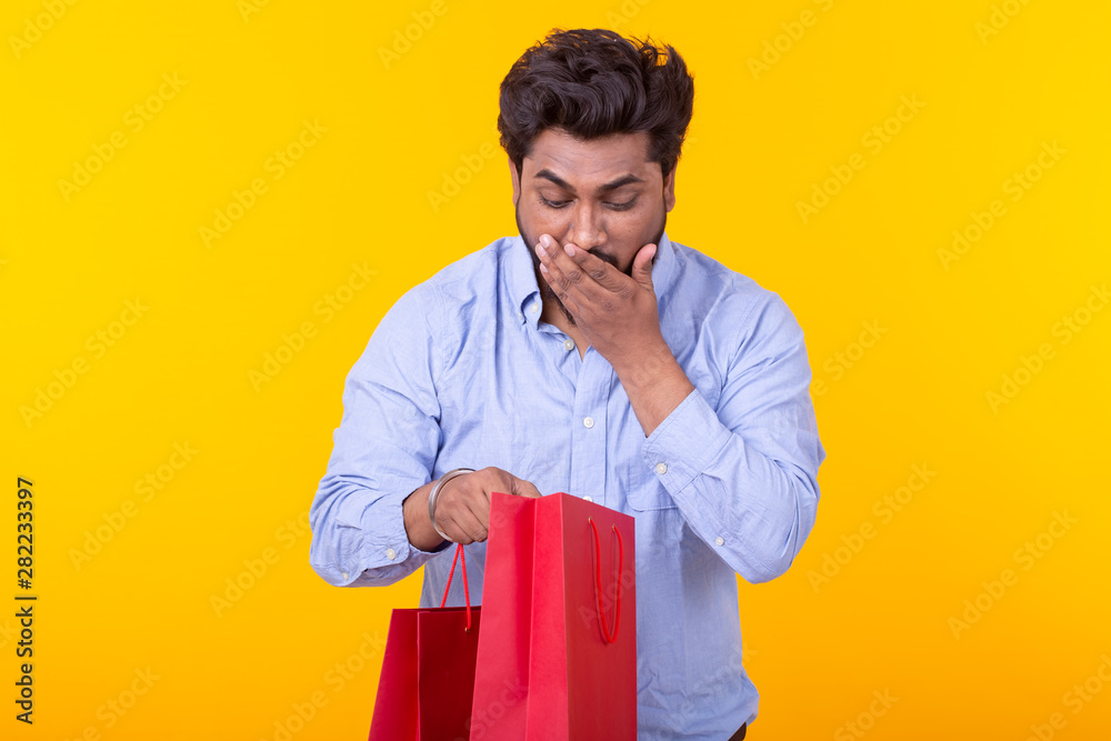 Surprised joyful young Indian man looks into the packages for shopping, standing on a yellow background. The concept of successful purchases. Surprised joyful young Indian man looks into the packages for shopping, standing on a yellow background. The concept of successful purchases.