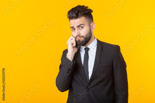 Cute young man with a beard in formal clothes talking on the phone posing on a yellow background. Concept of an informal meeting and business deal. © satura_