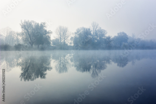 Morning hoarfrost on river. Autumn landscape with trees in fog reflecting in water. Natural background in cool blue colors