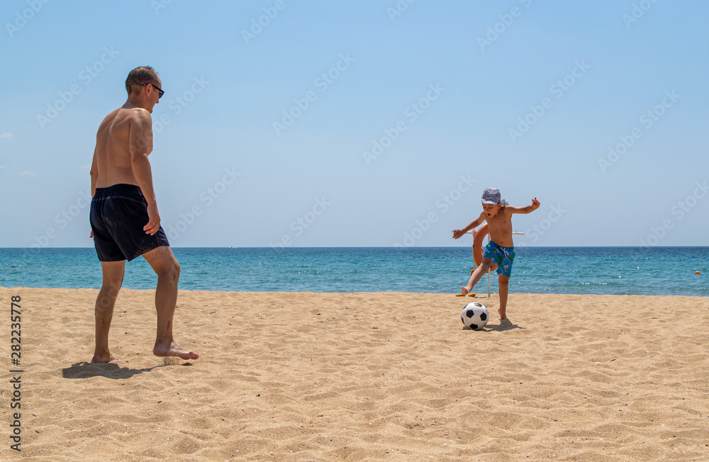 Father and son play football on the beach. The concept of sport and healthy lifestyle.Fresh sea air and good ecology. Friendship between father and son. Father and son play football on the beach. The concept of sport and healthy lifestyle.Fresh sea air and good ecology. Friendship between father and son.