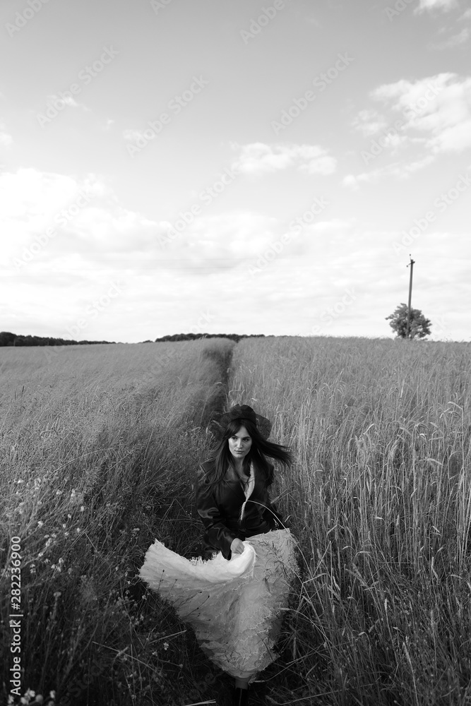 Black and white portrait of beautiful young woman in leather jacket, black hat, white dress., walking in grass.