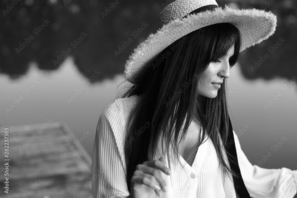 Black and white portrait of beautiful brunette woman in white shirt and big straw hat. Pond on background Black and white portrait of beautiful brunette woman in white shirt and big straw hat. Pond on background