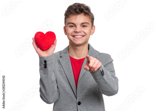 Happy Valentines Day. Cute young Teen Boy in Love with Red plush Heart in his hands. Smiling Child pointing finger to you and looking at camera, isolated on white background. © valiza14
