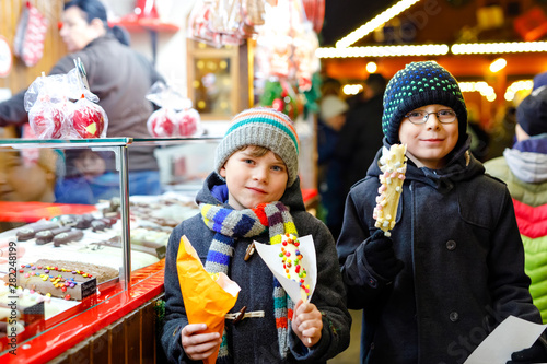 Two little kid boys, cute siblings eating bananas covered with chocolate, marshmellows and colorful sprinkles near sweet stand with gingerbread and nuts. Happy children on Christmas market in Germany.