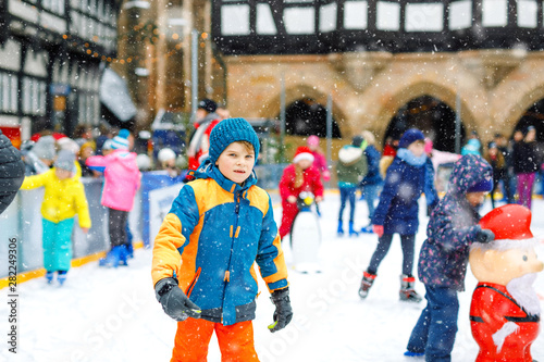 Happy little kid boy in colorful warm clothes skating on a rink of Christmas market or fair. Healthy child having fun on ice skate. Lot of people celebrating holiday and having active winter leisure