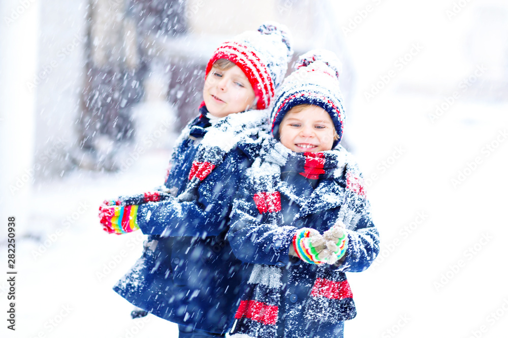 Two little kid boys in colorful fashion clothes playing outdoors during  strong snowfall. Active leisure with children in winter on cold days. Happy  siblings and twins having fun with first snow. Stock