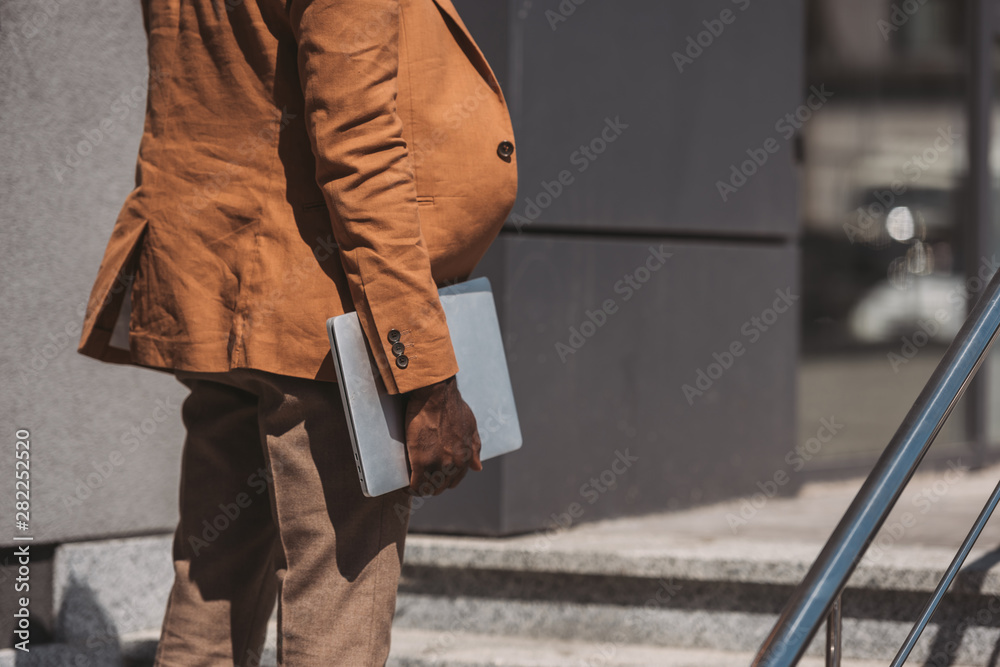 cropped view of african american businessman holding laptop while standing on stairs cropped view of african american businessman holding laptop while standing on stairs
