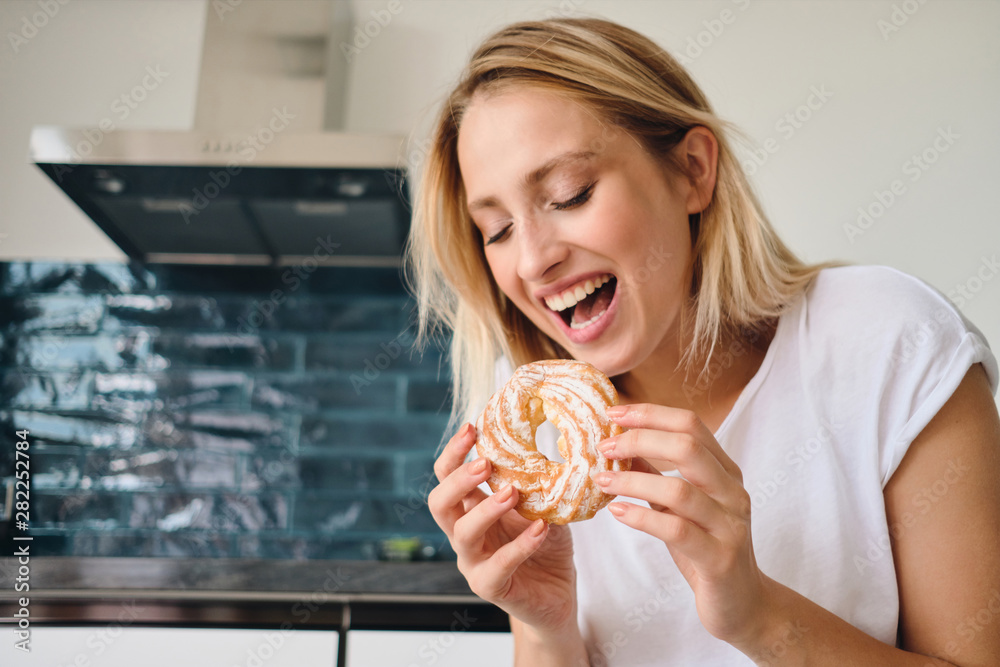 Young cheerful blond woman in white T-shirt emotionally eating sweet bun for breakfast at home Young cheerful blond woman in white T-shirt emotionally eating sweet bun for breakfast at home