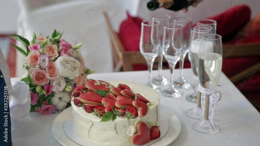 on the table are a cake with strawberries and heart cakes, a bride’s bouquet, champagne bottles in decorative packaging, the waiter pours champagne into glasses on the table are a cake with strawberries and heart cakes, a bride’s bouquet, champagne bottles in decorative packaging, the waiter pours champagne into glasses