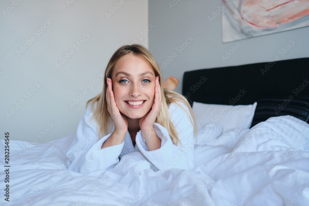 Young beautiful smiling blond woman in white bathrobe leaning on hands happily looking in camera lying in bed in modern hotel Young beautiful smiling blond woman in white bathrobe leaning on hands happily looking in camera lying in bed in modern hotel