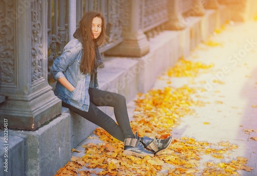Young pretty hipster woman sitting near iron fence in a trendy denim jacket black jeans and fashion trendy boots. Beautiful millenial girl and yellow golden autumn maple leaves. Fall on city street. © raisondtre