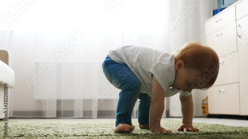 Baby girl falling down during learning to walk on floor in bright apartment.
