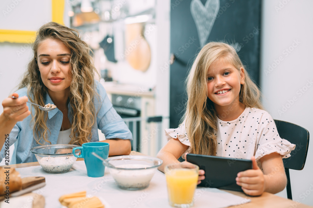 portrait of cute blond little girl using tablet while having breakfast with her mother at home