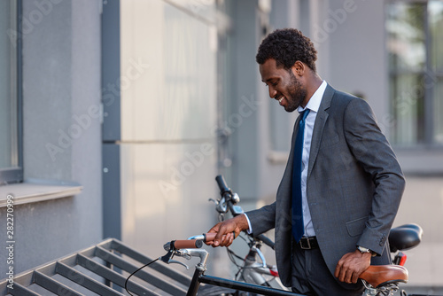 positive african american businessman smiling while taking bike from parking