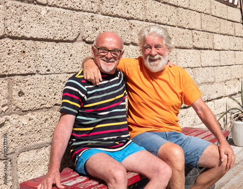 Two elderly Caucasian brothers hug while sitting on a wooden bench. Tropical place with bright sunshine. Bear and mustache for both people. Smiling looking at camera