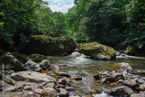 mountain river with large stones in the riverbed and stone banks, surrounded by forest along the banks, on a bright sunny day, with clouds in the sky.