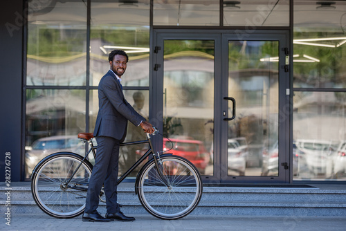 Fototapeta Naklejka Na Ścianę i Meble -  cheerful african american businessman smiling at camera while standing near office building