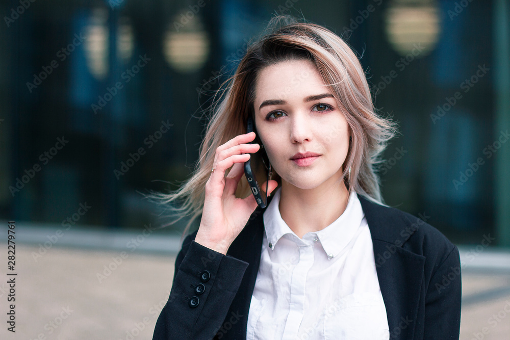 Businesswoman is talking by cell phone. Young formal dressed woman looking at camera. European girl outside work place in white shirt and black jacket. Office worker making a phone call. Business talk